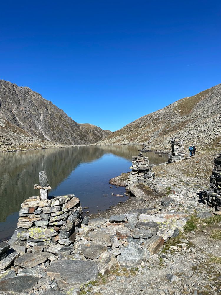 Im Bergsee spiegelt sichbder blaue Himme! Am Rande erkennt man dreinverschiedene rechteckige Gebilde aus vielen Steinen aufgetürmt