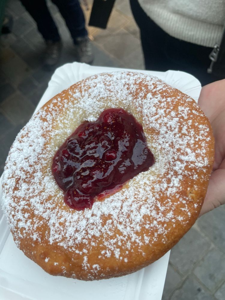 Das Bild zeigt ein warmes Kiachl – ein traditionelles, goldbraun ausgebackenes Tiroler Schmalzgebäck – das großzügig mit Staubzucker bestreut und in der Mitte mit einer Portion Preiselbeermarmelade gefüllt ist. Dieses Warme Kiachl wird auf einem weißen Pappteller serviert und vermittelt ein typisches, köstliches Markt- oder Weihnachtsmarkt-Erlebnis.