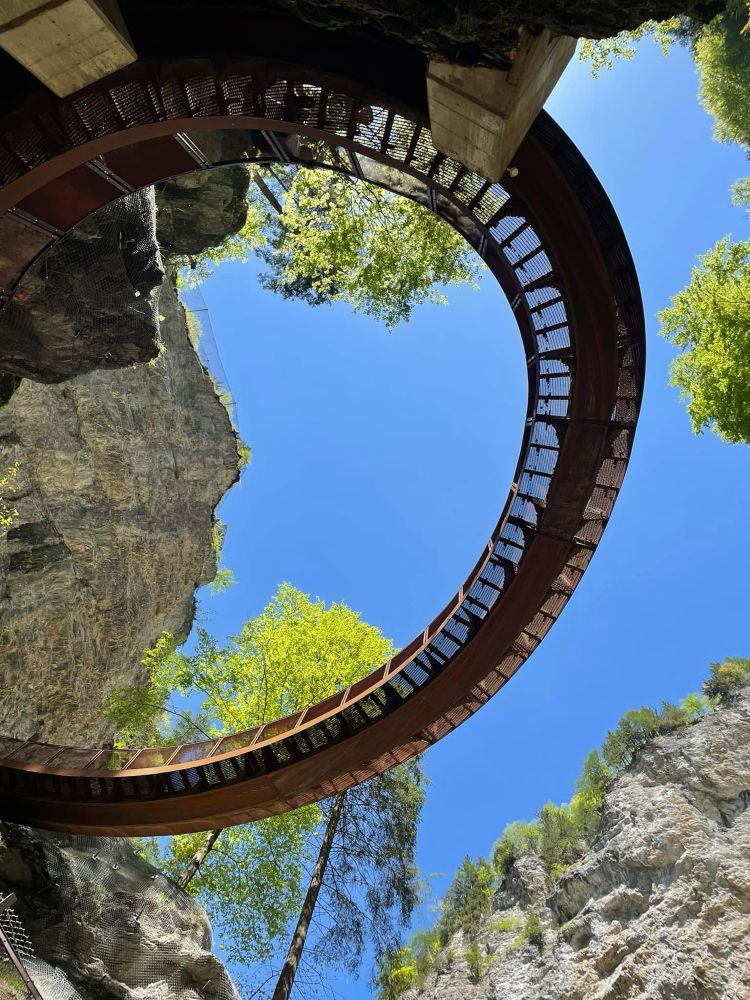 Man sieht einen Teil einer metallenen, rostroten Brücke, die sich im Bogen zwischen Felsen befindet! Fotografiert von unten gegen den blauen Himmel! Zwei leuchten grüne Laubbäume ragen dahinter empor!