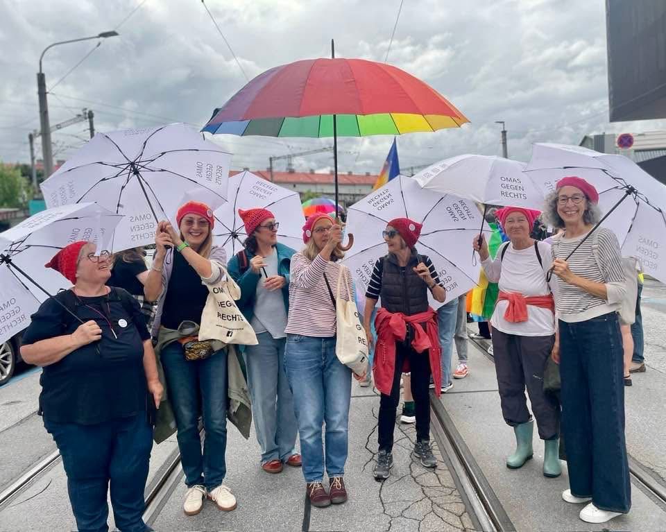 Das Bild zeigt eine fröhliche Gemeinschaft von Menschen, die mit weißen Regenschirmen und roten Mützen gemeinsam auf einer Straße stehen – ein starkes Symbol für Zusammenhalt, Solidarität und Engagement.