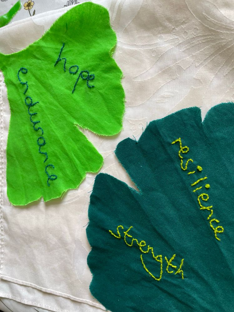 Two fabric pieces in the shape of ginkgo leaves, in two shades of green. The dark green leaf has the words resilience and strength embroidered on it. The bright green leaf has the words hope and endurance embroidered in it.