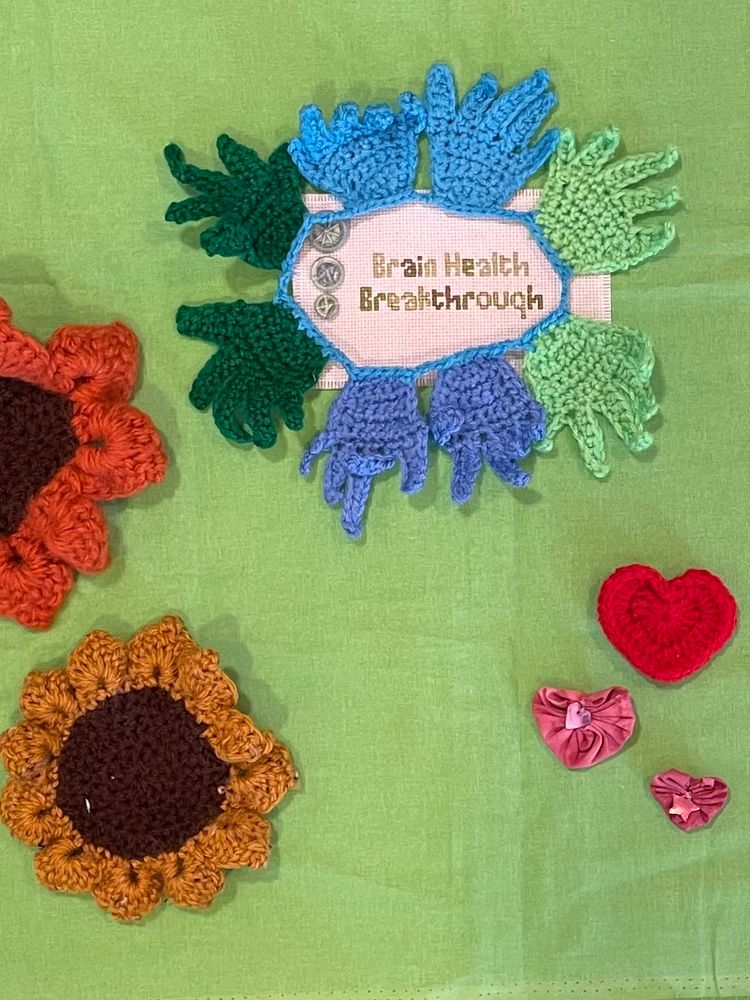 A selection of textile pieces laid out on a bright green background as part of a banner work in progress, including a cross stitch piece that reads ‘Brain Health Breakthrough’ circled by crocheted hands that are linked together. There are crochet sunflowers and a heart, and two pink Suffolk puffs in the shape of hearts.
