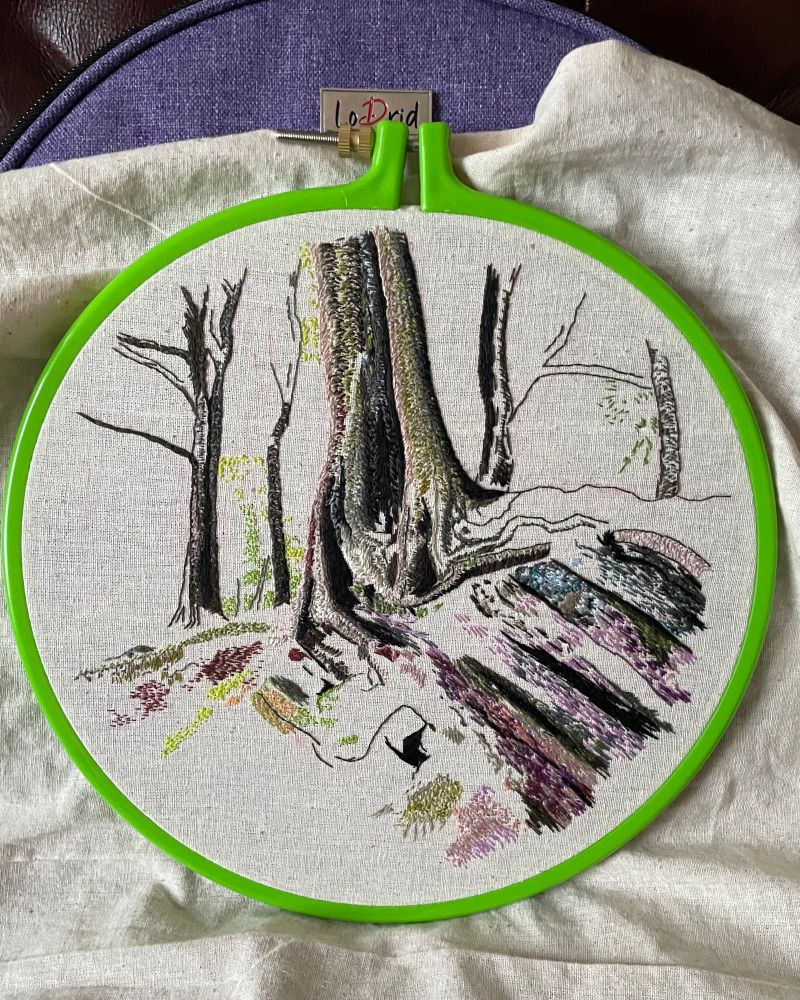 Hand embroidered ‘thread painting’ work in progress, depicting a woodland scene, with a central tree growing on rock, and framed in a green hoop.