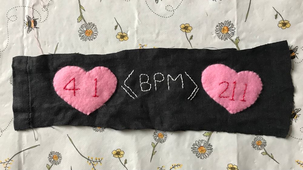 Textile piece. Black background fabric with two felt pink hearts. Stitched text goes across the hearts and background and reads 41<=BPM=>211