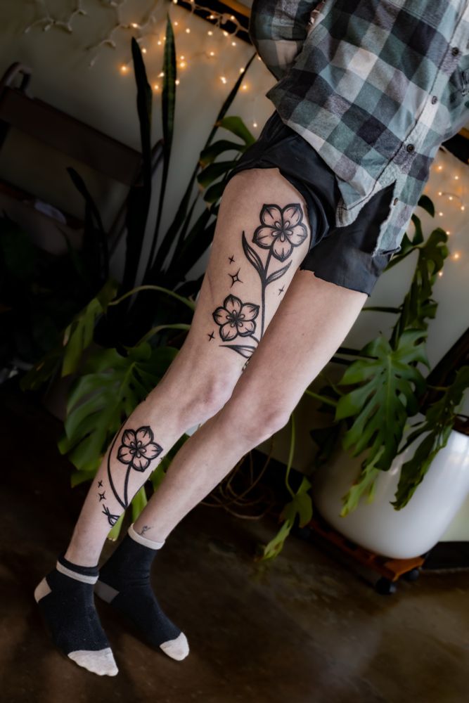 Photo of a person standing up, with a black tattoo of prairie gentian flowers wrapping around their right leg. 