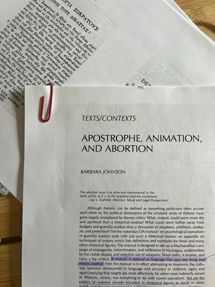 An image of an article that has been printed out (the pages are held together by a red paper clip in the top left corner and there is one sentence highlighted in purple). The article is Barbara Johnson’s “Apostrophe, Animation, and Abortion” 1986). It is set on top of another stack of papers (a printed version of the loeb of Xenophon’s Apology with only the Greek visible).