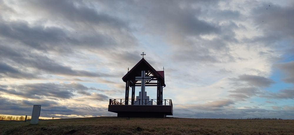 Paisaje de una pradera con una pequeña edificación. En su centro destaca una cruz blanca gigante. Sobre ella, hay un cielo cuyas nubes grisáceas están cortadas por el viento y en una esquina, a la izquierda, se puede apreciar como el atardecer dorado empieza a asomarse