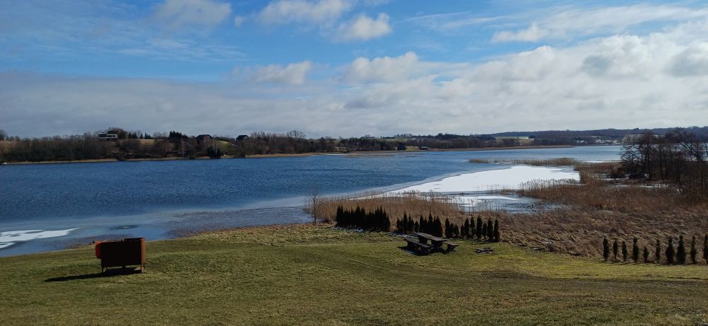 Foto de una pradera con un lago, también con parte de la costa congelada y un cielo azul con nubes blancas y grisáceas