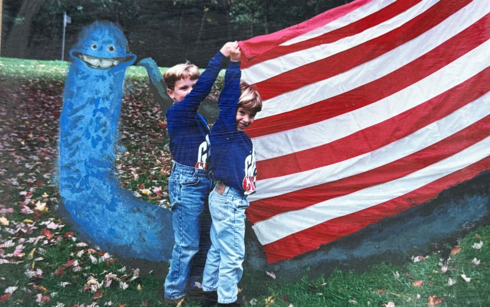 painted into a photograph of two children holding up an american flag, a long blue furry worm with a large smile creeps up behind them
