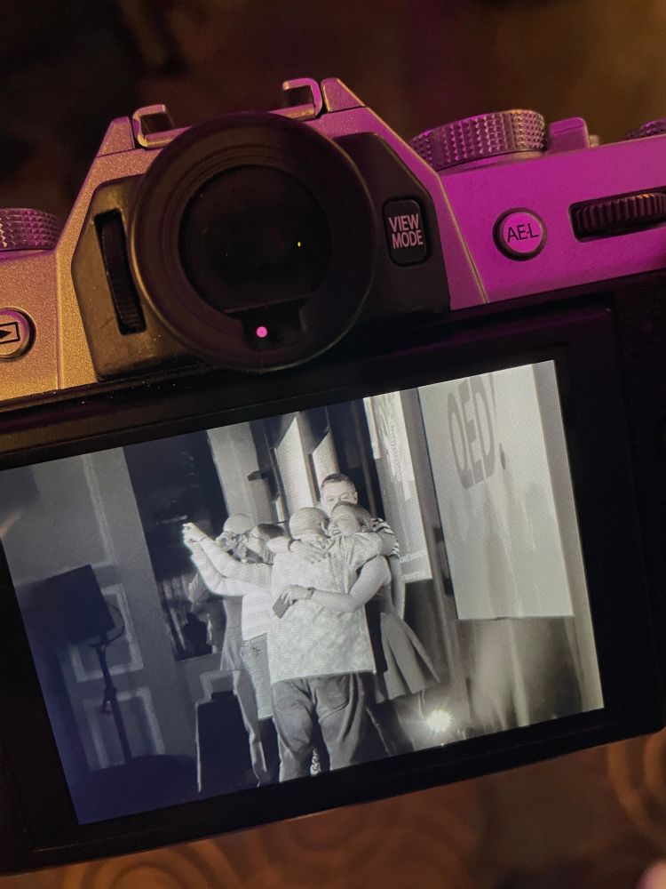 Shot of a camera’s rear screen showing a black-and-white photo of the QED organizers hugging on stage. 