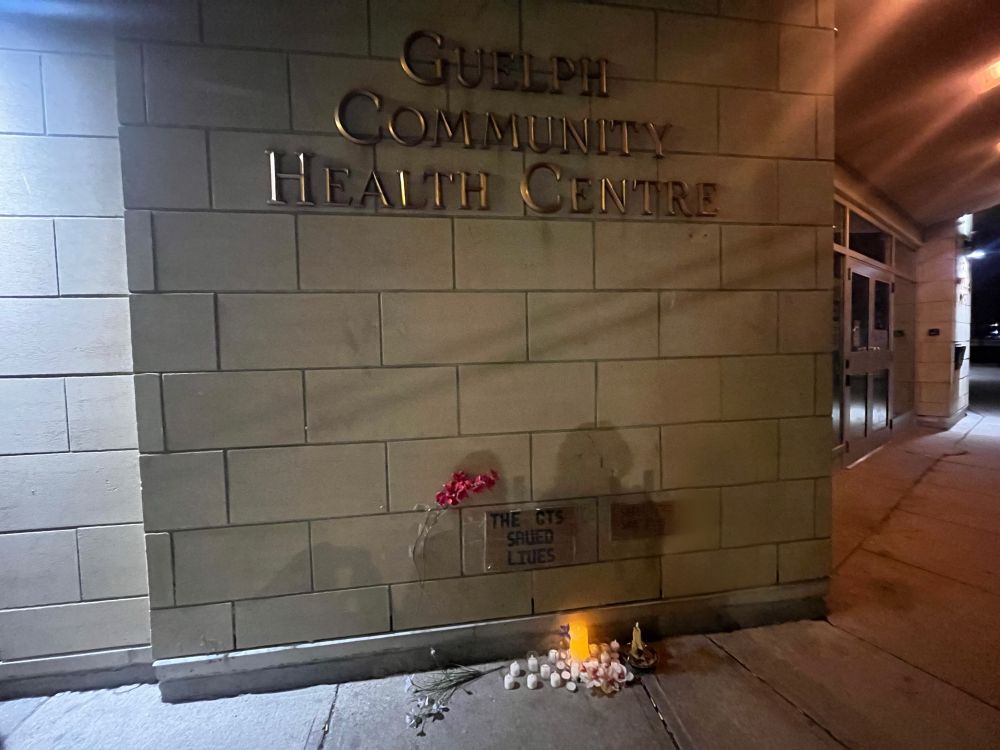 A small collection of candles, flowers, and naloxone kits placed along the edge of a sidewalk next to a building under two cardboard signs. One sign reads “THE CTS SAVED LIVES” the other reads “SAFE SITES SAVE LIVES”. A permanent sign on the side of the building reads “Guelph Community Health Centre”