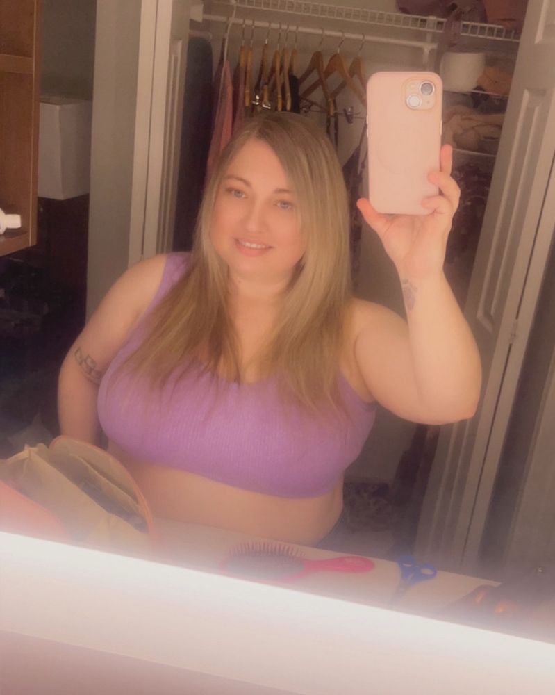 A mirror selfie of Lorelei in a lavender crop top with side bangs. The bathroom counter in front of her has a pink hairbrush, a blue pair of scissors, a makeup bag, and a hair straightener