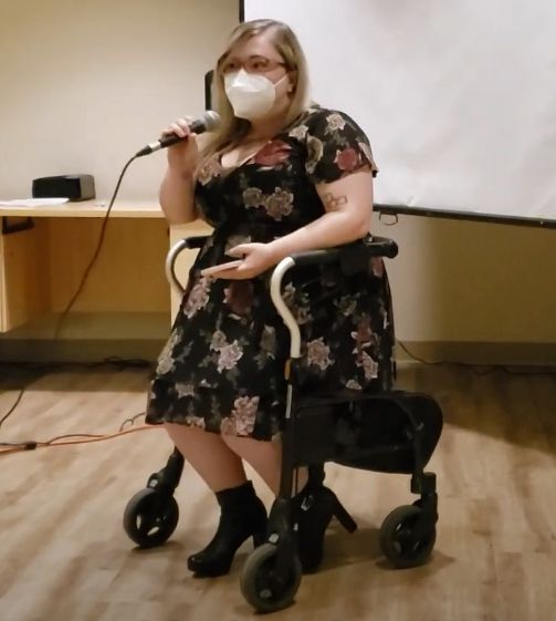 Lorelei in a black floral dress with black boots sitting on her walker. She is wearing a white respirator. She is presenting in front of a projector screen and holding a microphone