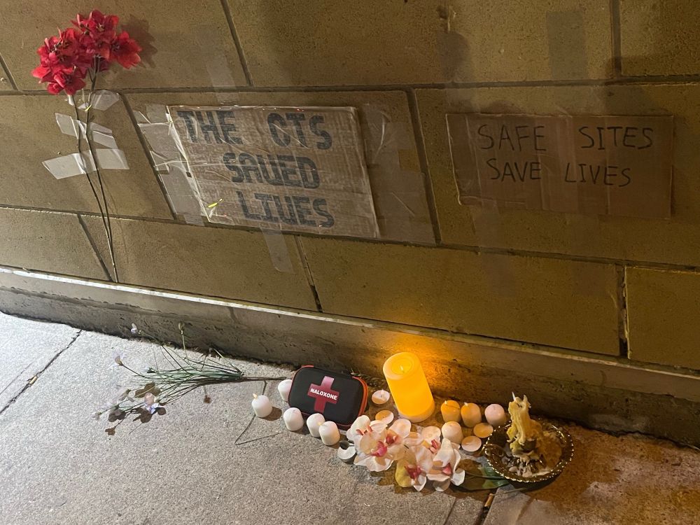 A small collection of candles, flowers, and naloxone kits placed along the edge of a sidewalk next to a building under two cardboard signs. One sign reads “THE CTS SAVED LIVES” the other reads “SAFE SITES SAVE LIVES”
