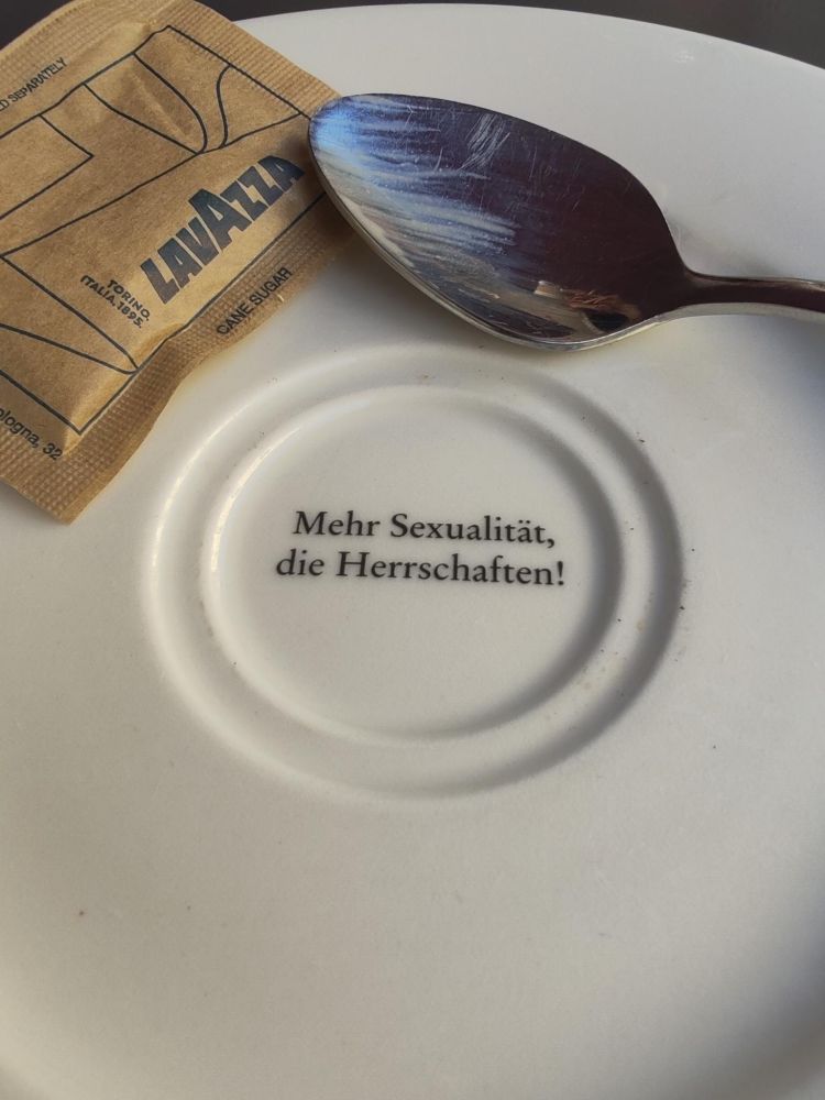Auf einem weißen Teller liegt ein silberner Löffel, der teilweise auf dem Teller ruht. Neben dem Löffel befindet sich ein braunes Papiertütchen mit der Aufschrift 'LAVAZZA'. In der Mitte des Tellers steht der Text 'Mehr Sexualität, die Herrschaften!' in schwarzer Schrift.