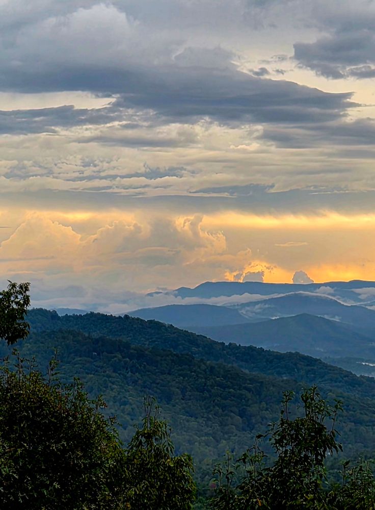 Yellow strip of sky, grey and white clouds, mountains still deep green.