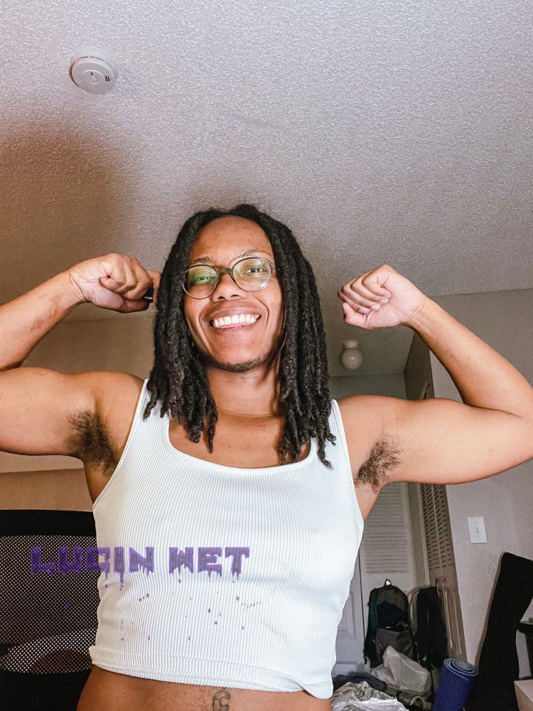 Lucin Wet smiling at the camera as they flex their biceps, wearing a cropped white tank top. 