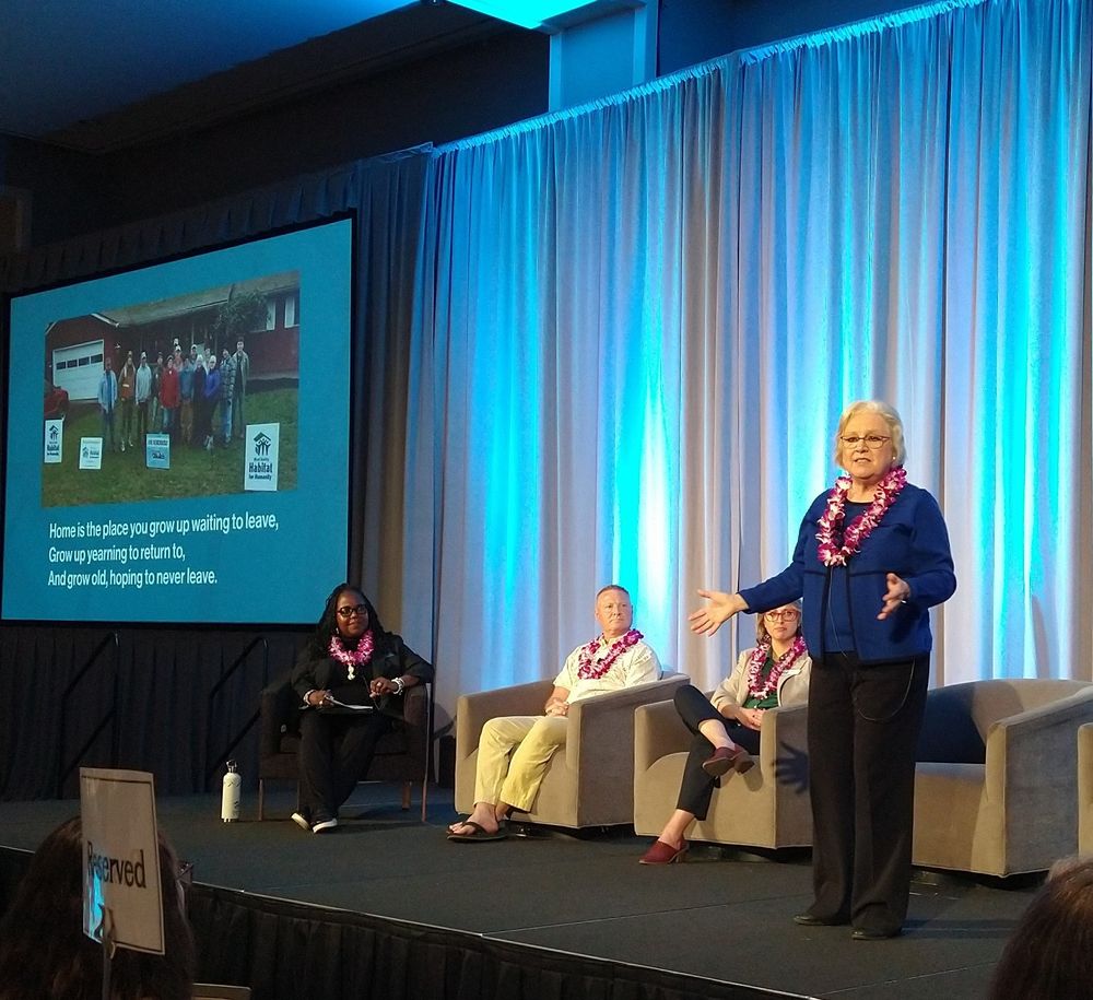 A woman stands on a stage, talking to an unseen audience, while three other people sit on the stage listening to her. There is a slide show behind her. 