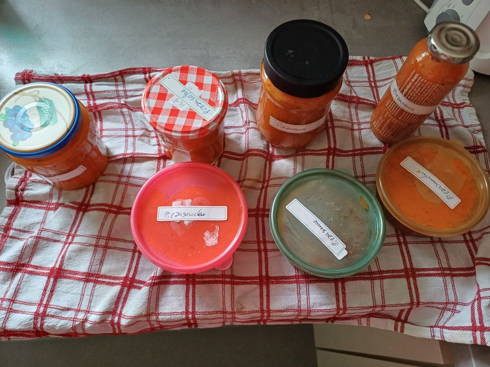 Ein rot-weiß kariertes Küchenhandtuch. Darauf 4 verschiedene Schraubgläser und 3 runde, bunte Plastikdosen gefüllt mit roter Tomatensauce.  