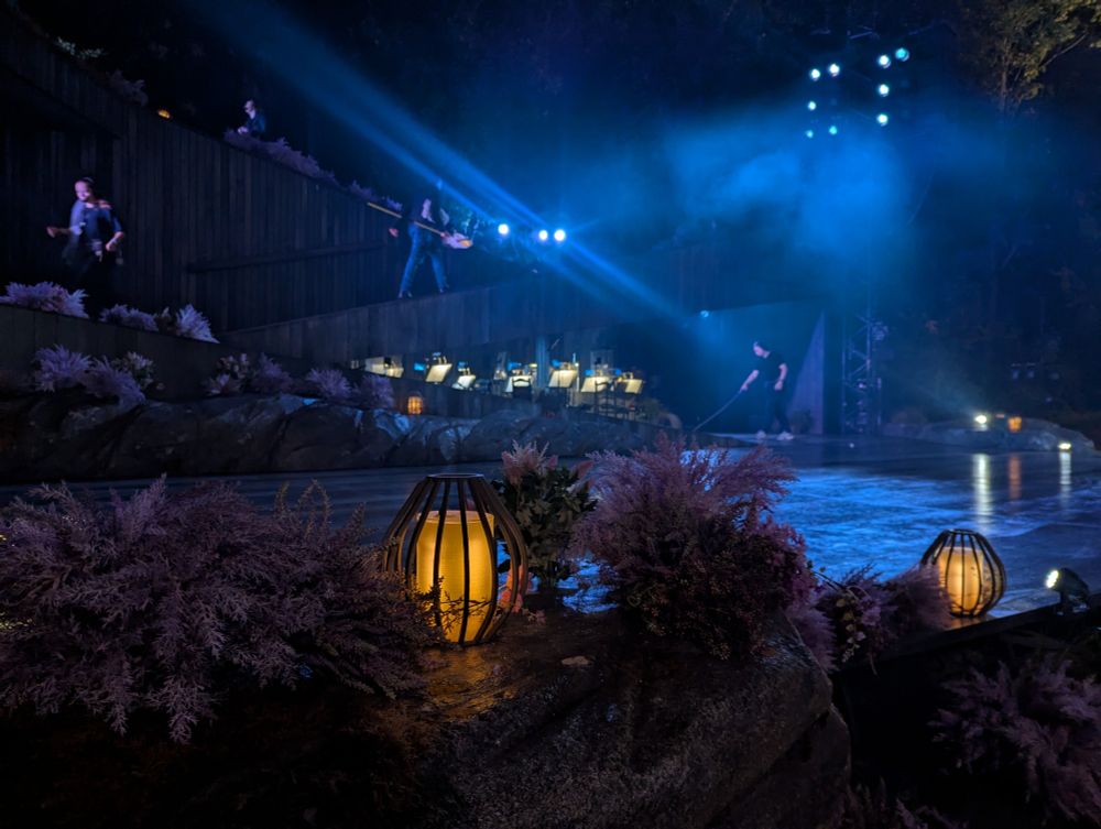 夜の屋外ステージで青い照明に照らされた演出準備中の様子。前景にランタンと紫のアザミ、背景に演奏者と作業員が見える。