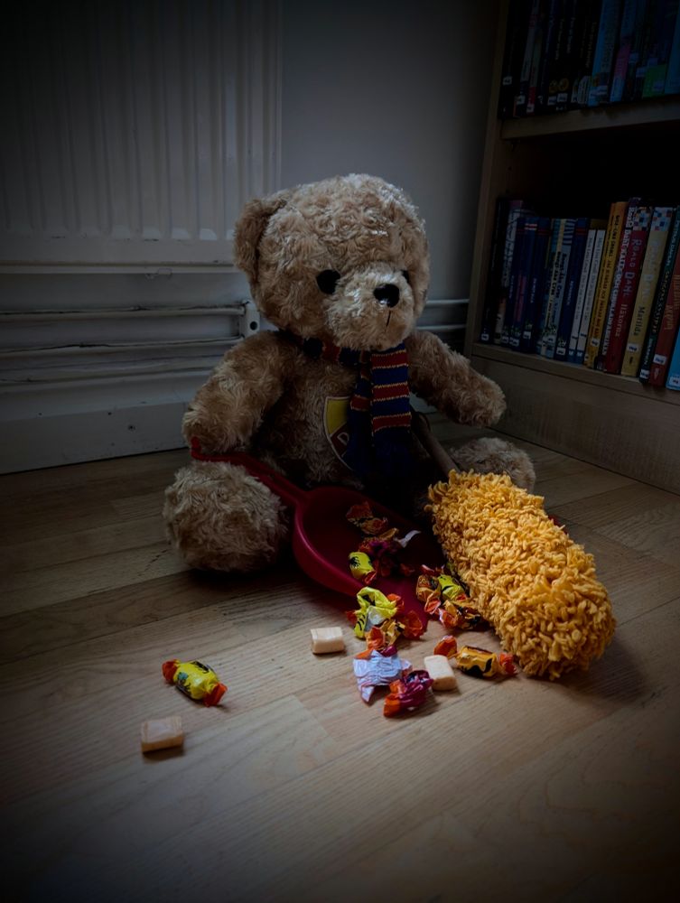 En nallebjörn sitter på golvet och skyfflar upp godis och godispapper. I bakgrunden böcker. 
