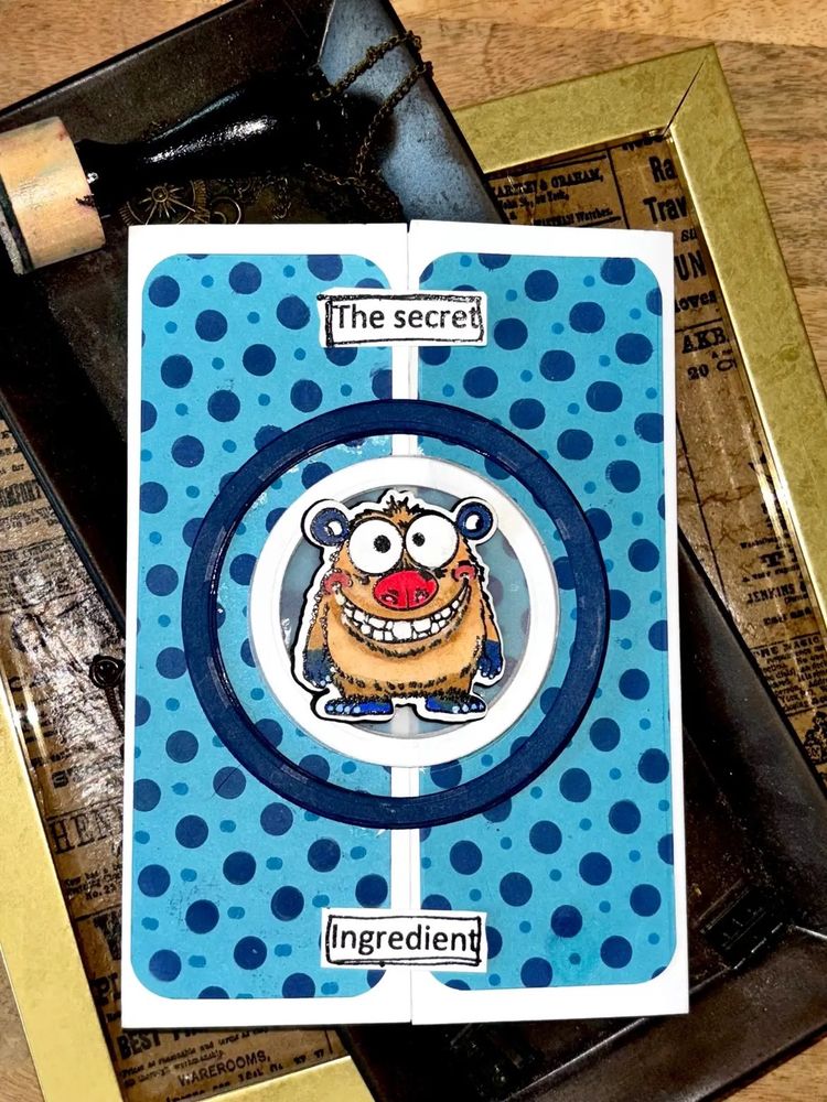 Cut paper and stamp-ink art blue polka dot card. At the center is a wild eye, square toothed, squat bear. The words “the secret ingredient” also frame the figure. 