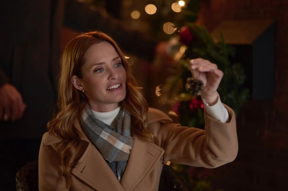 A young white woman with strawberry blonde hair and a camel trench coat holds up a pinecone Christmas tree ornament which she looks at with wonder.