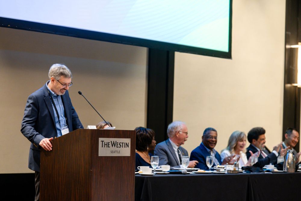 John Budd speaking at a podium during the 77th Annual Meeting of the Labor and Employment Relations Association. Members of the head table next to the podium are laughing. 