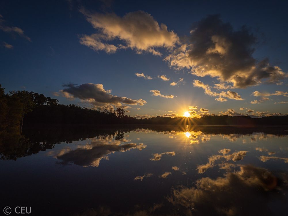 The sun rises over the still surface of a lake, its rays breaking through scattered clouds. Thick cloud groupings reflect on the surface, doubling the sense of depth and light and pulling in the viewer. 