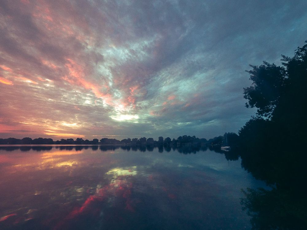 An expansive lake scene just prior to sunrise, with dramatic cloud cover lit in pastel shades - yellows, pinks, powder blue. The colorful sky is reflected nearly exactly in the calm water below. Trees line the distant horizon, and a small sailboat sits quietly near the wooded shore on the right.