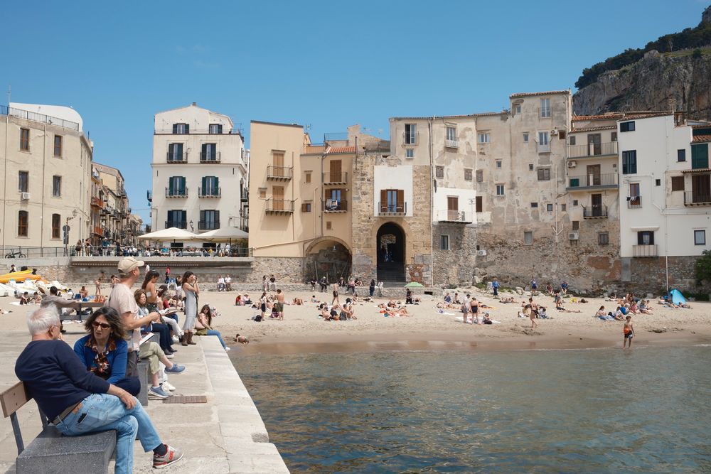 Crowded Cefalu beach with historic buildings. 