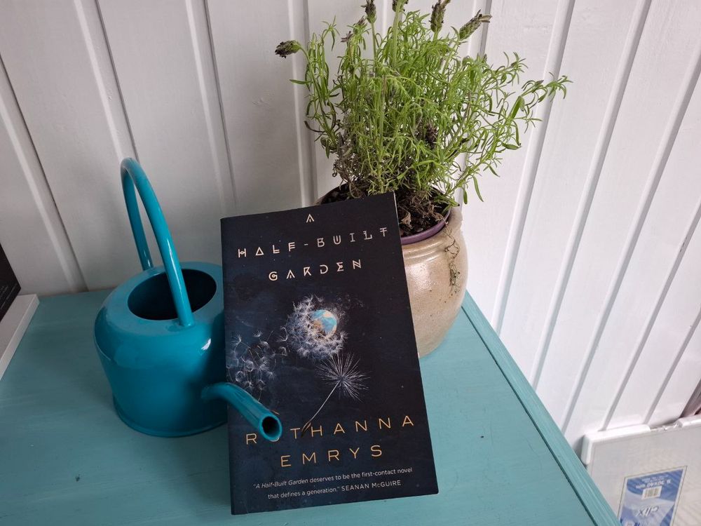 Das Buch "A Half-Built Garden" von Ruthanna Emrys steht an einen Topf mit Lavendel und eine Gießkanne gelehnt auf einer Kommode vor einer weißen Holzwand.