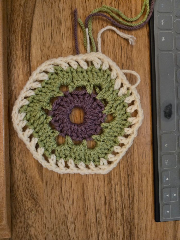 Crochet in purple, green and cream layers, shaped like a hexagon.