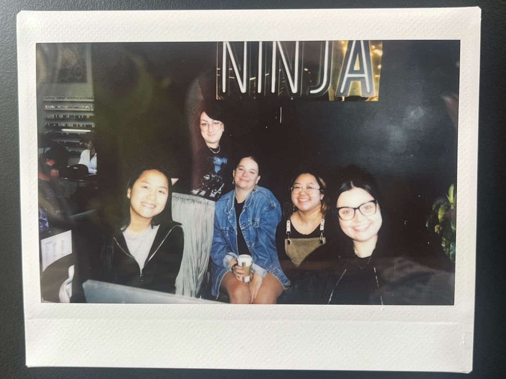 a photo of a polaroid of five people behind the front desk of a nail salon, posing and smiling together