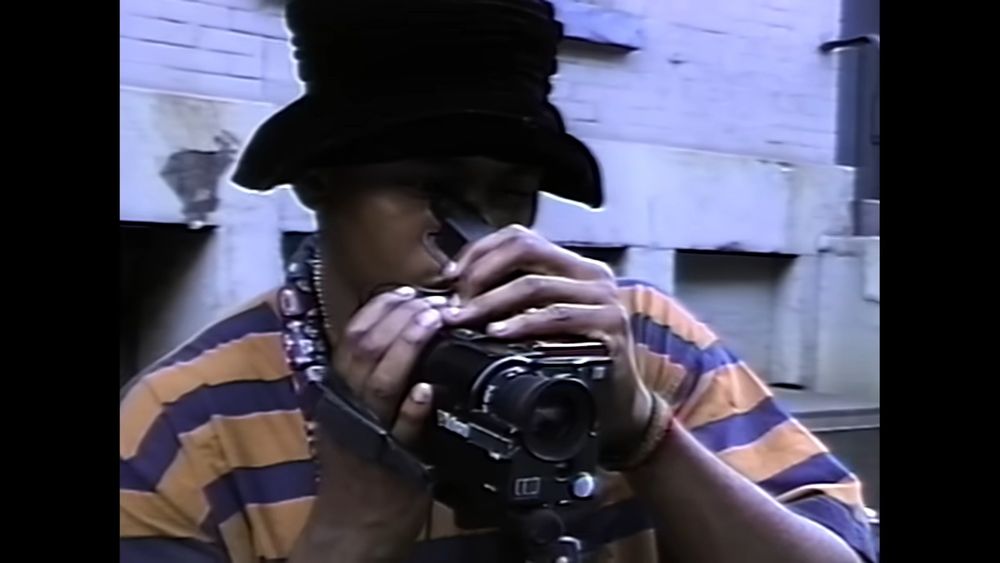 LL Cool J in a striped purple and yellow shirt and a large black hat using a camcorder.