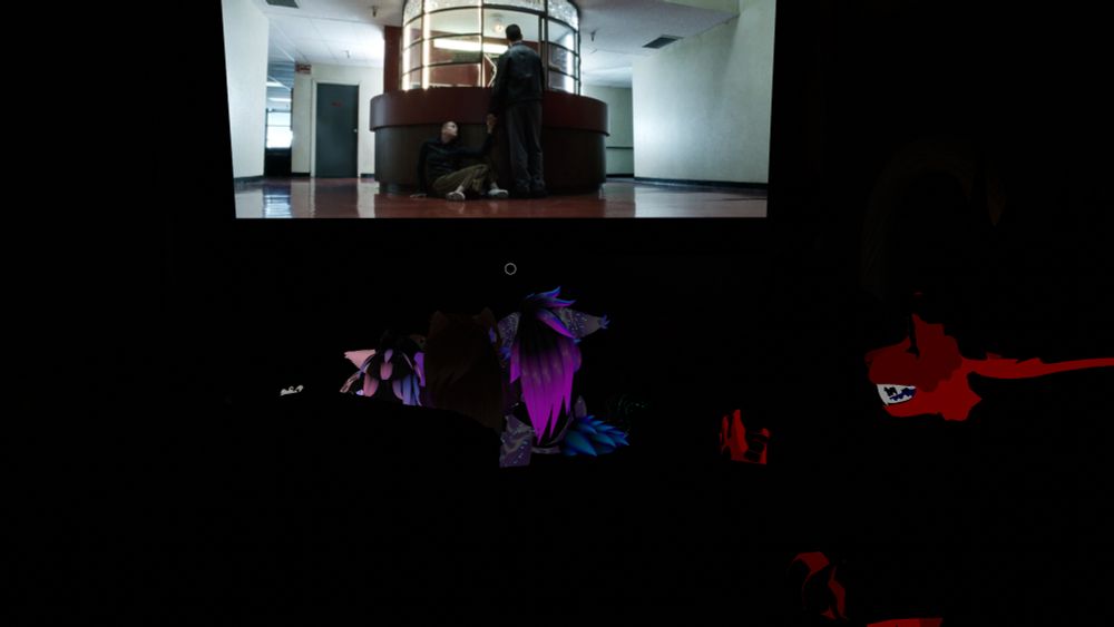 A dark theater-like room with a large screen showing a movie scene. On the screen, a man stands beside a circular reception desk while another person sits on the floor leaning against it. The hallway around them is brightly lit with plain walls and a reflective floor. In the foreground, silhouetted humanoid and creature-like avatars are faintly visible watching the screen.