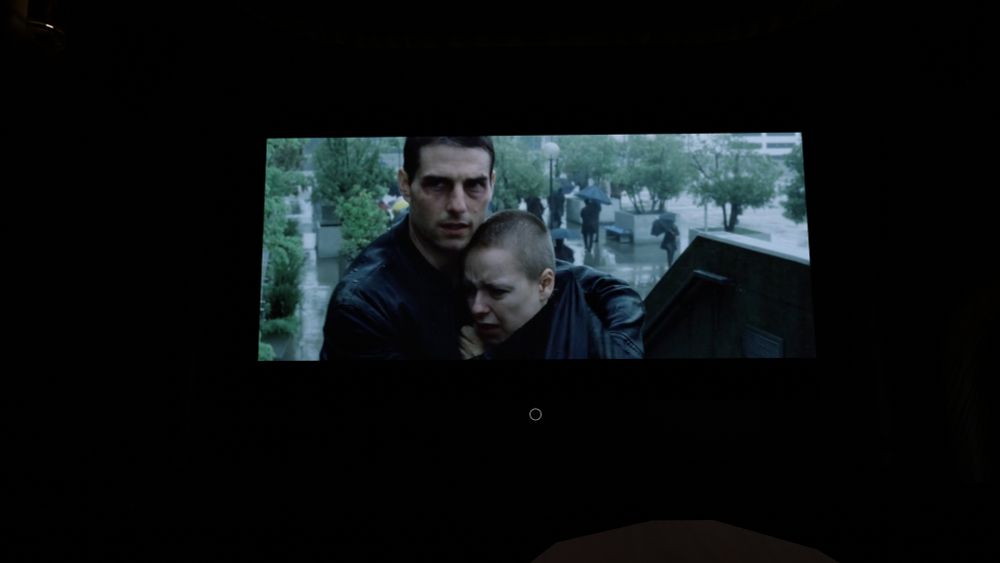A dark room with a large screen showing a movie scene. On the screen, a man in a dark jacket is holding and protecting a distressed person with a shaved head. It is raining heavily in the background, with umbrellas and people walking in a city plaza. The rest of the room is almost completely dark except for the illuminated screen.