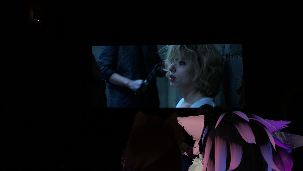 A dark theater interior shows a large screen with a scene of a blonde woman sitting against a wall, lit in cool blue tones, while several brightly colored VRChat avatars watch from the seats in front.