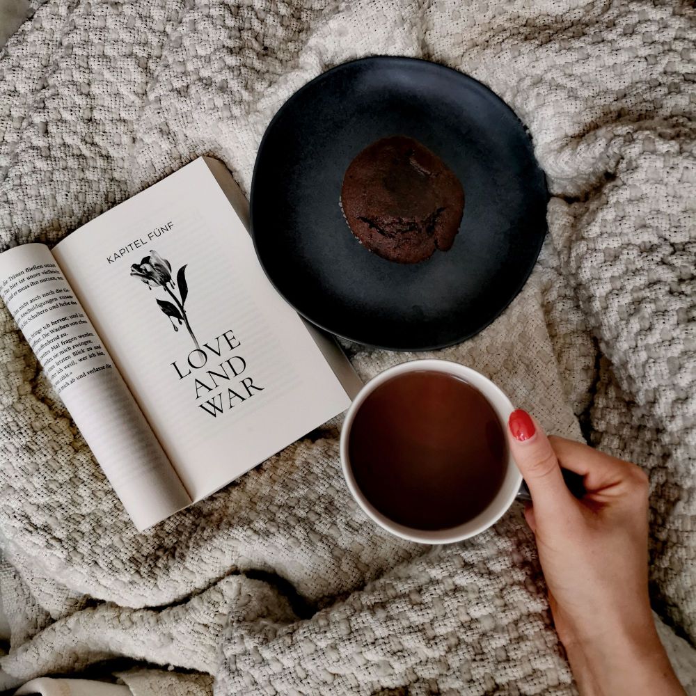 Ein aufgeschlagenes Buch auf einer beigen Decke. Daneben ein Teller mit Muffin und eine, von einer Hand gehaltenen, Tasse mit Tee.