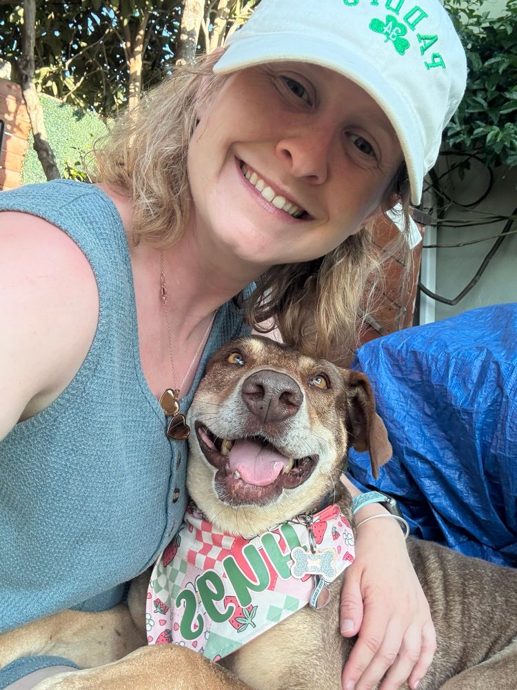 Hugs, the brown dog with floppy ears, looks up at Rachel, his foster-turned-forever human, lovingly. He is sporting a colorful bandana with his name on it and everyone is smiling.