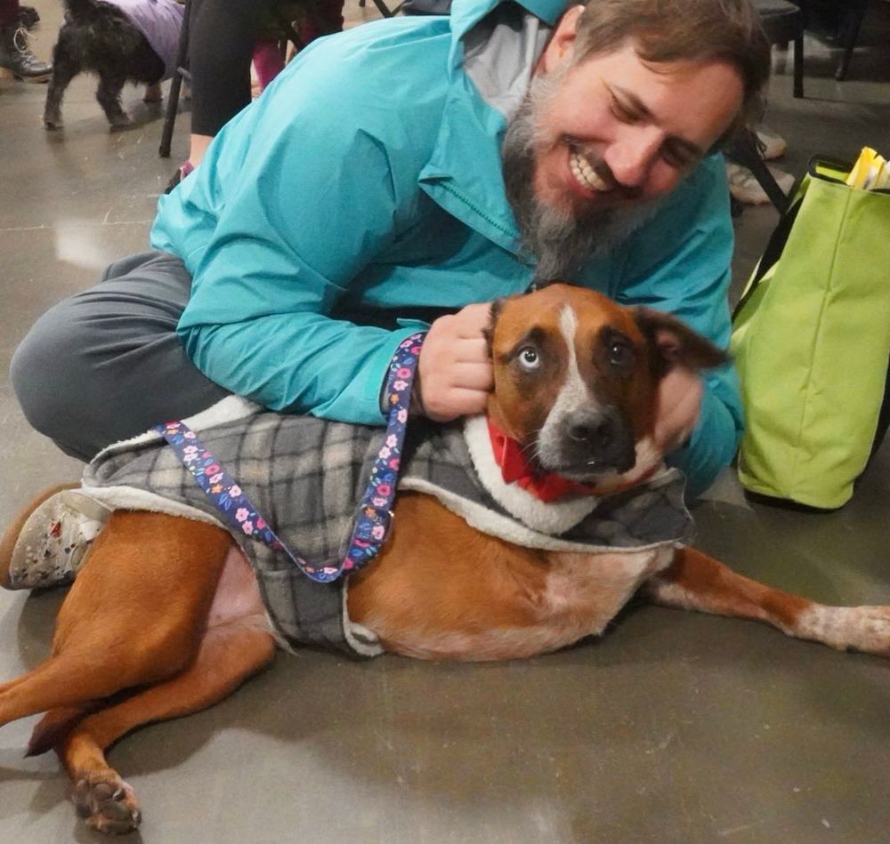 Jennie is wrapped in a plaid coat at a bustling adoption event, lying flat with her legs stretched out as her new forever human embraces her from behind.