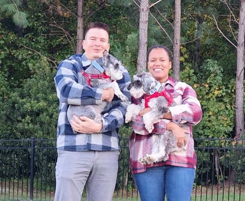 sarge poses with his new forever family, which includes a doggy sibling named Enzo who looks exactly like him. Each parent holds up one pup and it’s a beautiful day.