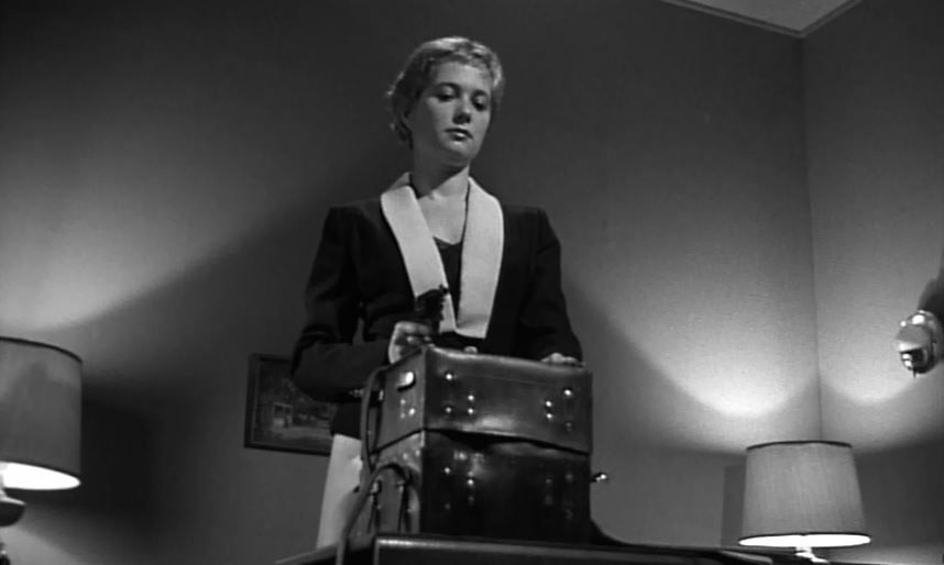 B&W still of a woman holding a gun and looking down at a bulky, hinged box.