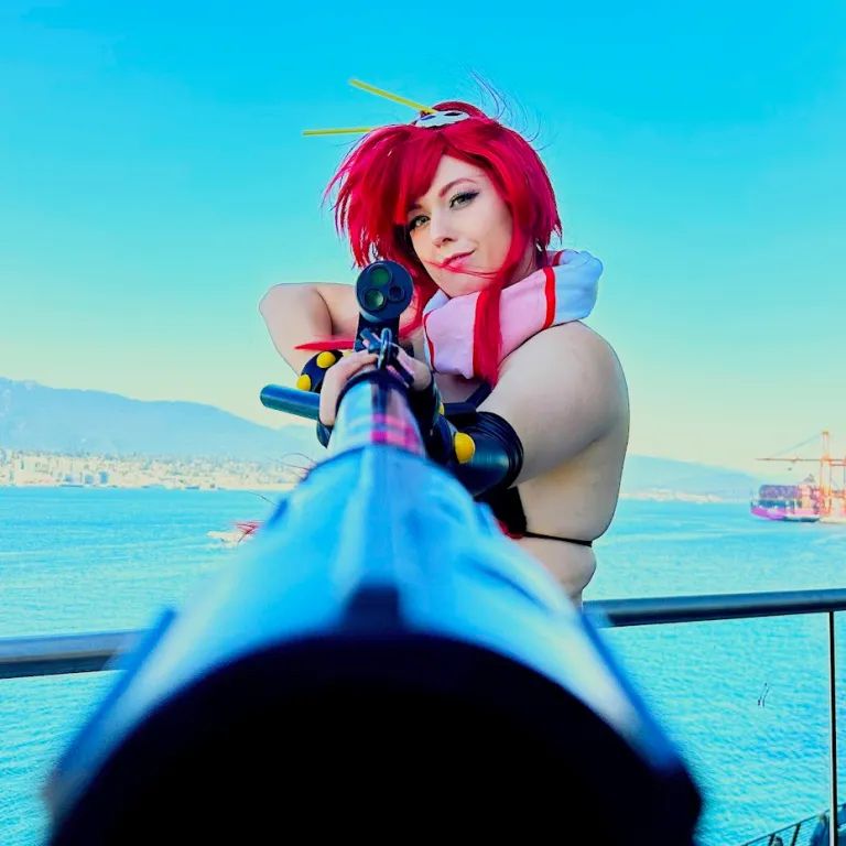 A photograph of Cosplaimee. She is looking down the barrel of a prop firearm, with the ocean in the background.