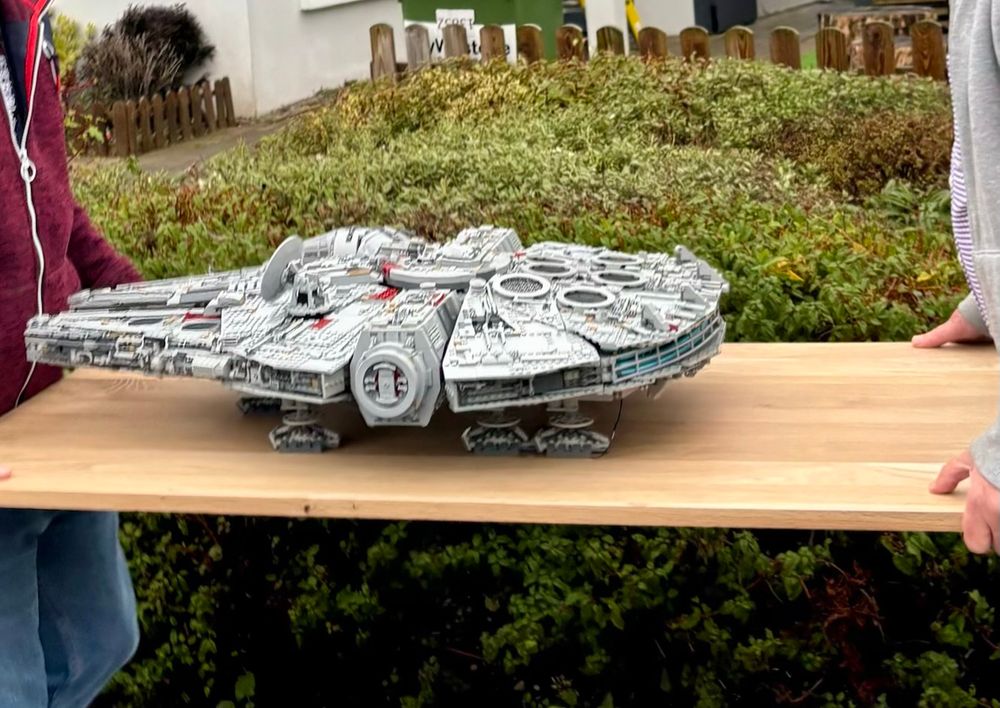 A big-ass LEGO Millenium Falcon on a board being carried through a suburban neighbourhood, currently passing in front of an exuberant hedge.