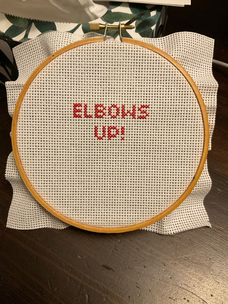 The words “Elbows Up!” cross stitched in red block letters on a white background. The fabric is held by a wooden embroidery hoop. There is enough space for an angry Canadian to stitch a maple leaf under the letters and put a border around it all, once she figures out what she wants it to look like.