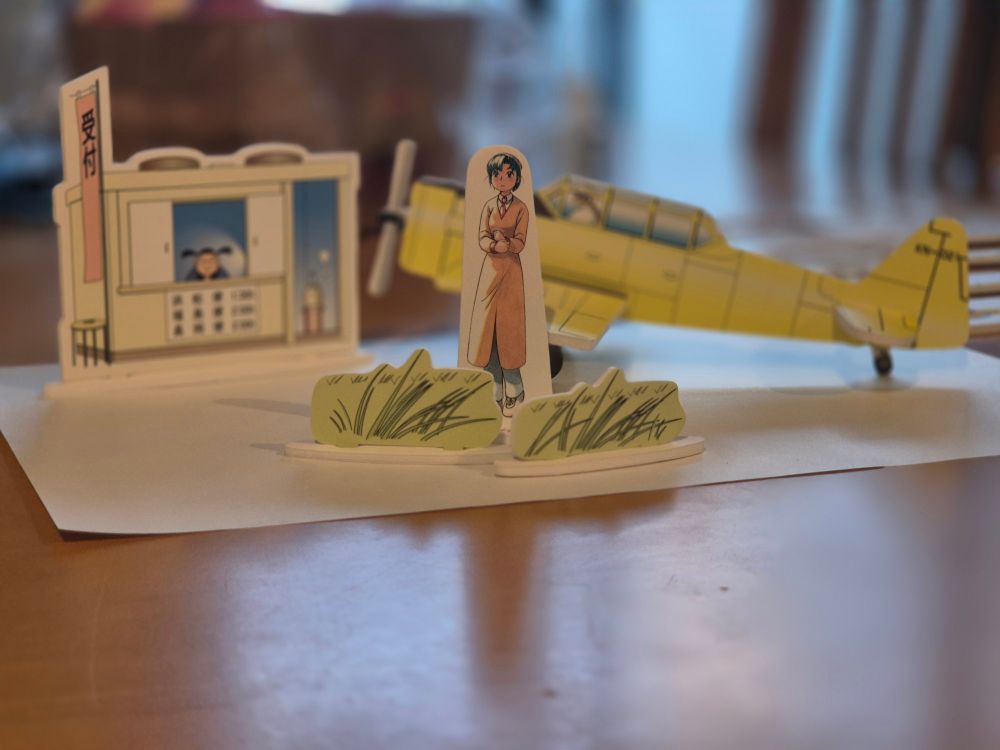 Alpha from YKK next to a shop and a plane (cardboard and paper, 2025)