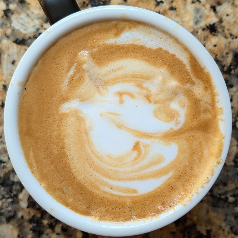 A cup of latte with foam art resembling a cartoon-style dog face. The design shows a rounded muzzle, a pointed ear, and a curved line suggesting an eye, surrounded by swirls of brown crema on the white foam.