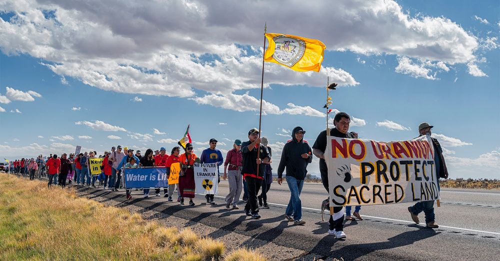 Image of a line of people walking along the side of a road with signs to protest a uranium mill.