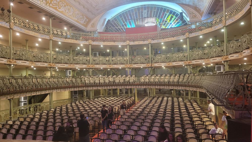 Foto do Teatro José de Alencar. Embaixo cadeiras em formato antigo. Três andares de camarotes. Em cima vitrais coloridos. 